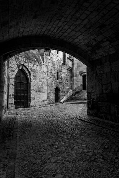 A black and white photograph showing an old cobblestone street. The closest section is covered, while the back section is open, but ends in a dead end with a door.