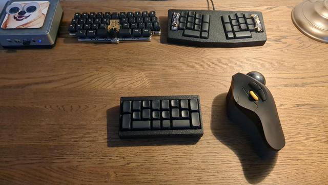 A small keyboard on a wooden desk next to a larger trackball mouse. Two additional keyboards and a mini PCin the background.
