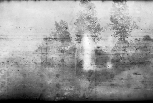 Very old black and white photo. Foggy and spotted, with markings from the backing paper showing, you can barely make out some trees and a person in old fashioned clothes.