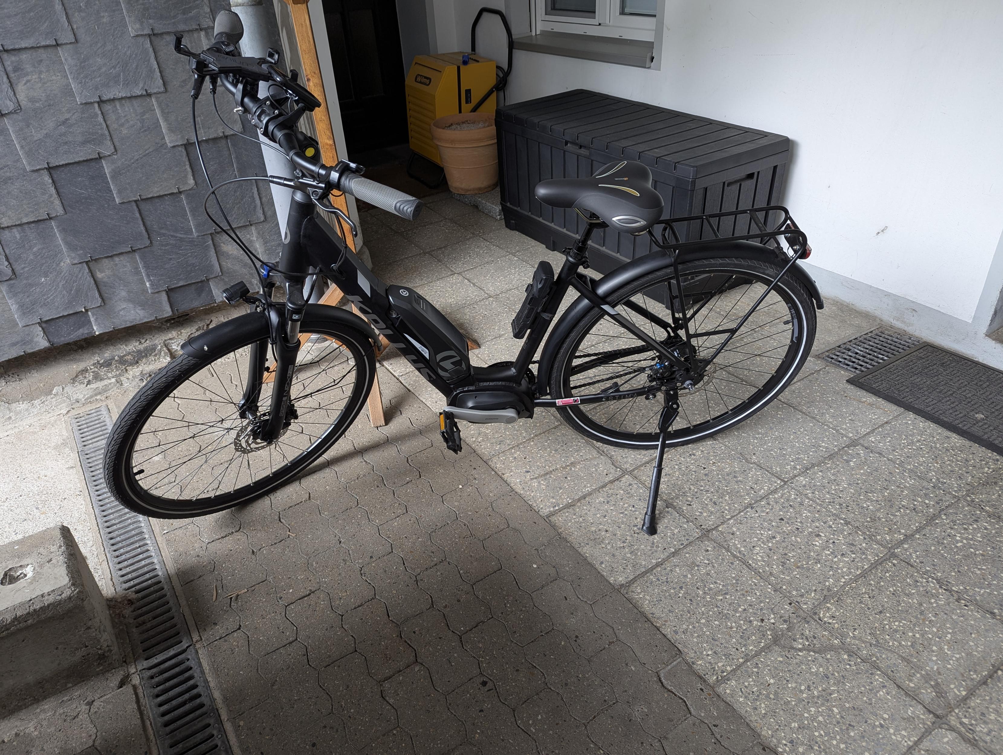Ein schwarzes E-Bike auf einem gepflasterten Hof, die Wand links hinter dem Rad ist mit Schiefer verkleidet, die rechts weiß gestrichen. An der Wand steht eine schwarze Box für Gartenmöbel Auflagen.