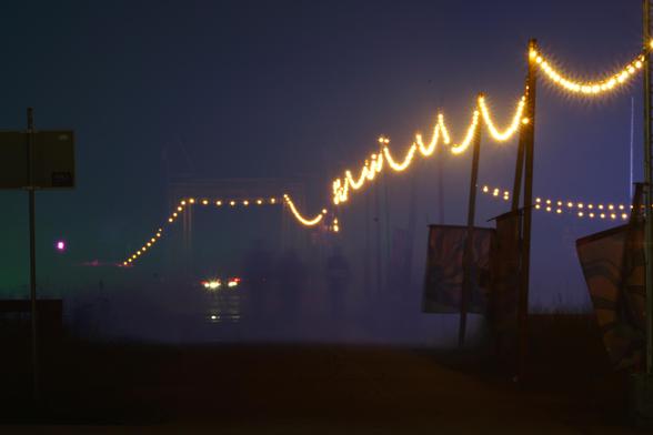 A foggy night with strings of warm lights spanning along a street. Some shadows of people in the distance emerge from the fog.