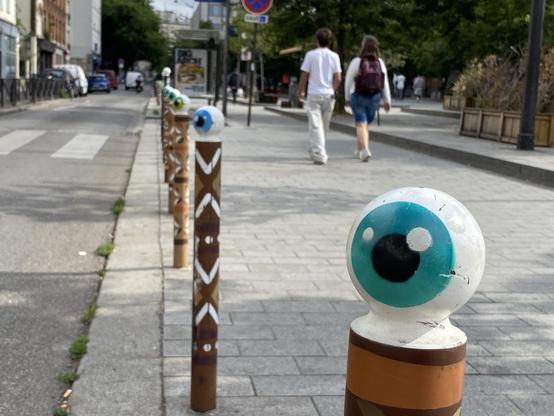 Straßenszene mit einer Reihe von Pollern. Auf der runden Endungen ist ein Auge aufgemalt.