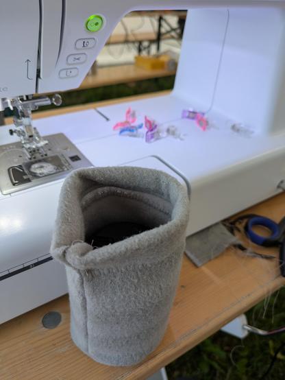 An empty gray bag standing up. In the background is part of a sewing machine and some utensils visible.