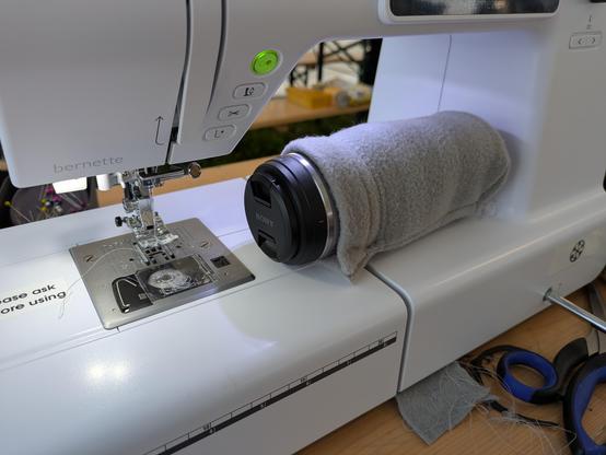 A camera lens half covered in a nearly finished lens case. It is laying on a sewing machine on its side.