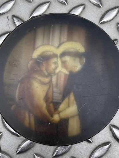 Ein kreisförmiges Bild im Vintage-Stil, das zwei Figuren zeigt, deren Stirnen sich berühren und die sich an den Händen halten, vor einem weichen Hintergrund. Die Figuren scheinen historische oder traditionelle Kleidung zu tragen. Das Bild wird von einem dunklen, kreisförmigen Rahmen umrahmt und befindet sich auf einem strukturierten