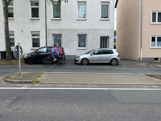 Ein Radfahrer mit einem lila Kapuzenpulli fährt neben zwei auf einem Gehweg an einer Querungshilfe geparkten Autos - einem schwarzen und einem silbernen - eine Straße entlang. Im Hintergrund sind zwei Wohngebäude mit mehreren Fenstern zu sehen. Auf der linken Seite ist ein Straßenschild zu sehen.
