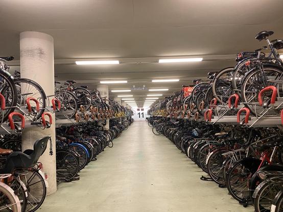 Ein Fahrradparkhaus mit mehreren Etagen von Fahrradständern. Zahlreiche Fahrräder sind ordentlich auf jeder Seite untergebracht, so dass ein langer schmaler Gang entsteht, der zu einem sichtbaren Ausgang am anderen Ende führt. Helle Oberlichter erhellen den Raum.