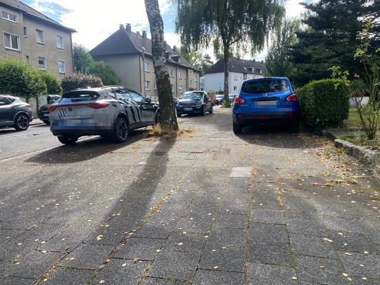 Das Bild zeigt eine Straßenszene mit geparkten Autos, in zwei Reihe auf einem Gehweg. Im Hintergrund befinden sich Wohngebäude, Bäume an der Seite und ein gepflasterter Gehweg mit verstreutem Laub.