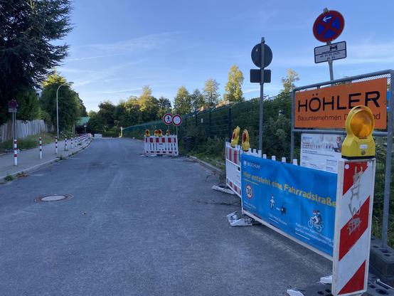 Eine Sackgasse mit angepolterten Gehweg, Baustellenabsperrungen und einem Banner auf dem steht hier entsteht eine Fahrradstraße, darunter einige Symbole, wie auf der Fahrradstraße, die Verkehrsteilnehmer:innen sich verhalten sollen.