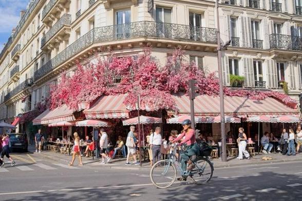 Rosa Blumen an einem Cafe mit rosa Markise. Eine Radfahrende Person mit rosa Oberteil quert diese Szene