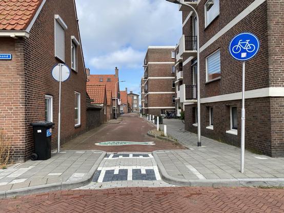 Eine ruhige Wohnstraße mit Backsteinhäusern und Ziegeldächern. Es gibt Straßenschilder für Radfahrer, eine Mülltonne und eine Kreuzung mit Kopfsteinpflaster. Die Szene ist teilweise bewölkt und zeigt eine Mischung aus Backstein und Beton in einer städtischen Umgebung.