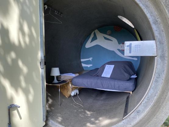 Ein einzigartiger Schlafplatz in einem großen Betonrohr. Im Inneren befinden sich ein Bett mit grauer Bettwäsche, ein kleiner Nachttisch mit einer Lampe und ein buntes Wandgemälde an der Wand. An der Außenseite ist ein Tastenschloss zu sehen.