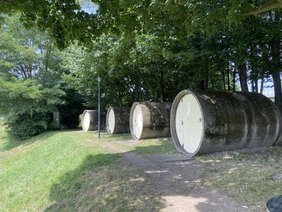 Das Bild zeigt drei große, zylindrische Betonstrukturen, die auf einer Grasfläche unter Bäumen stehen. Jeder Zylinder hat eine weiße Tür und wirkt verwittert. Ein Weg führt zwischen den Strukturen hindurch, die von etwas Grün umgeben sind.