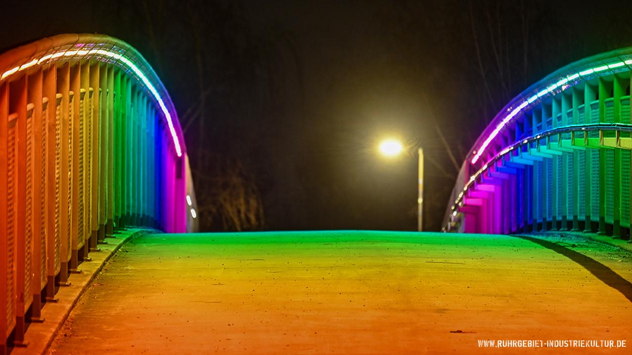 Nächtliche Aufnahme einer beleuchteten Fußgängerbrücke mit regenbogenfarbener LED-Beleuchtung. Die Brücke hat Geländer auf beiden Seiten, die in einem fließenden Farbverlauf von Orange und Gelb über Grün und Blau bis hin zu Violett und Pink illuminiert sind. Der Gehweg selbst wird durch das bunte Licht in warmen Gelb- und Grüntönen getaucht. Im Hintergrund ist eine Straßenlaterne sichtbar. Nächtliche Aufnahme einer beleuchteten Fußgängerbrücke mit regenbogenfarbener LED-Beleuchtung. Die Brücke hat Geländer auf beiden Seiten, die in einem fließenden Farbverlauf von Orange und Gelb über Grün und Blau bis hin zu Violett und Pink illuminiert sind. Der Gehweg selbst wird durch das bunte Licht in warmen Gelb- und Grüntönen getaucht. Im Hintergrund ist eine Straßenlaterne sichtbar.