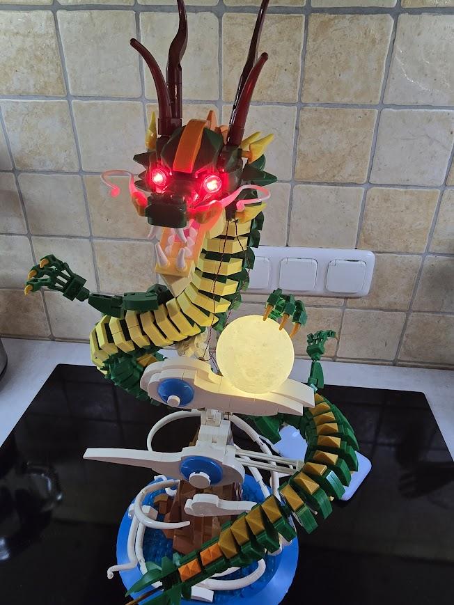 Ein chinesischer Drache aus Klemmbausteinen. Der Drache scheint in Wolken zu schweben und eine seiner Klauen "greift" nach einer runden Kugel, die wie der Mond aussieht. Das Modell steht vor gekachelter Wand auf einem schwarzen Kochfeld, ist etwa 50 cm hoch, die Augen des Drachen leuchten rot, der "Mond" leuchtet gelbweiß. Ein chinesischer Drache aus Klemmbausteinen. Der Drache scheint in Wolken zu schweben und eine seiner Klauen "greift" nach einer runden Kugel, die wie der Mond aussieht. Das Modell steht vor gekachelter Wand auf einem schwarzen Kochfeld, ist etwa 50 cm hoch, die Augen des Drachen leuchten rot, der "Mond" leuchtet gelbweiß.