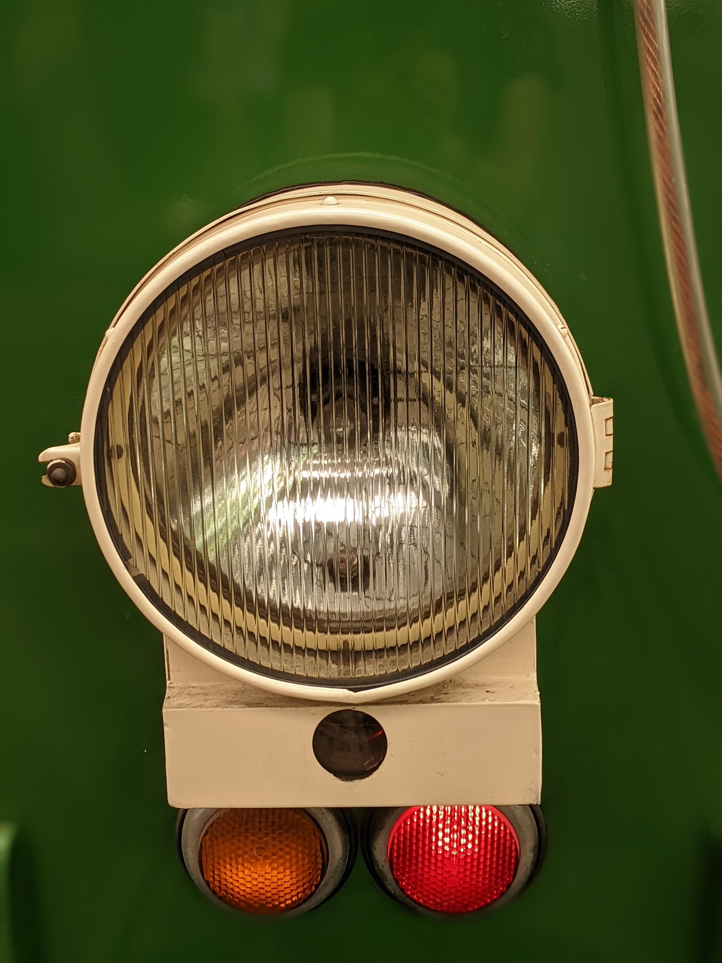Ein Frontscheinwerfer an einem grünen Fahrzeug. Der runde Scheinwerfer hat ein vertikal geriffeltes Glas und eine weiße Einfassung. Darunter befinden sich zwei kleinere, runde Lichter: ein orangefarbenes auf der linken und ein rotes auf der rechten Seite. Das gesamte Licht ist an einem grünen Hintergrund montiert. Ein Frontscheinwerfer an einem grünen Fahrzeug. Der runde Scheinwerfer hat ein vertikal geriffeltes Glas und eine weiße Einfassung. Darunter befinden sich zwei kleinere, runde Lichter: ein orangefarbenes auf der linken und ein rotes auf der rechten Seite. Das gesamte Licht ist an einem grünen Hintergrund montiert.