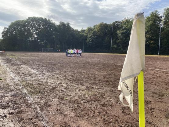 Ein schlammiges Fußballfeld mit einer Eckfahne im Vordergrund. Im Hintergrund ist eine Gruppe von Spielern versammelt, die wahrscheinlich Strategien besprechen, während Bäume den Rand des Feldes säumen und der Himmel bewölkt ist.