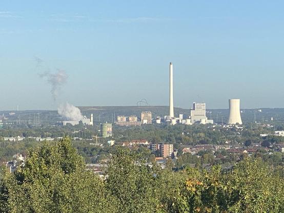 Ein Blick auf ein Industriegebiet mit mehreren Fabriken und Kühltürmen, die Dampf ausstoßen, vor einem klaren blauen Himmel und umgeben von Grün.
Im Hintergrund eine Halde mit einer Stahlkonstruktion. 