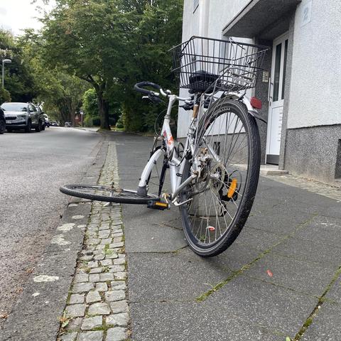Ein silbernes Fahrrad mit einem vorderen Korb lehnt an einem Pfosten. Das Vorderrad ist demontiert und liegt neben dem Rad. 
