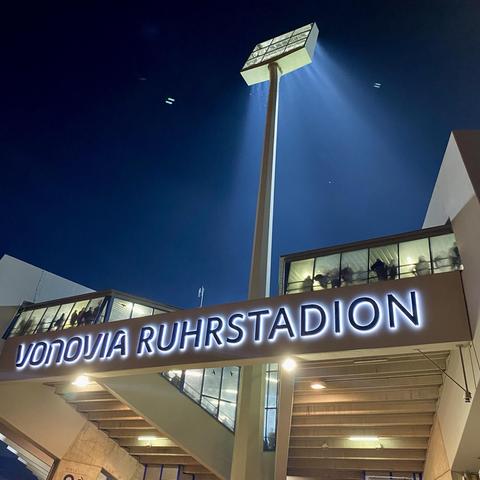 Das Bild zeigt den Eingang des Vonovia Ruhrstadions bei Nacht, mit beleuchteter Beschilderung und einem hohen Scheinwerfer, der Lichtstrahlen wirft. Im Hintergrund sind die Silhouetten von Menschen durch Glasscheiben auf einem Balkon zu sehen.