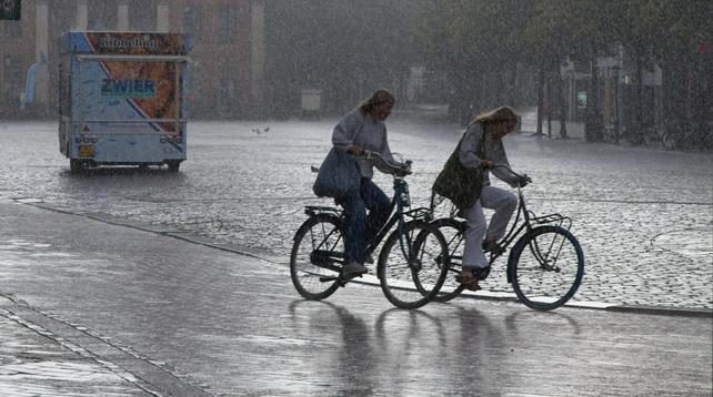 zwei Radfahrende Personen bei Regen  in einer Stadt. Im Hintergrund ein gepflasterter großer Platz.