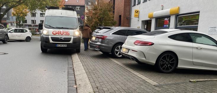 Ein Lieferwagen ist in der Nähe des Bordsteins geparkt, während eine Person daneben geht. Mehrere Autos, darunter ein grauer Geländewagen und ein weißes Sportcoupé, sind dicht nebeneinander auf der Straße geparkt.