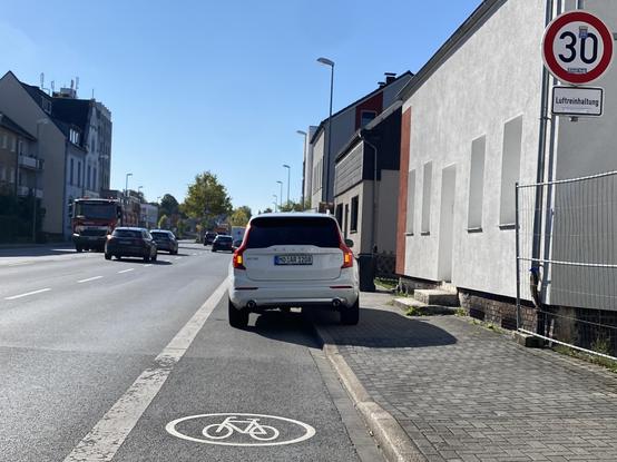 Ein Auto parkt halb auf einem Gehweg und halb auf einem Radweg. Wenige Meter vor dem Auto ist deutlich ein Fahrrad-Piktogramm zu sehen.