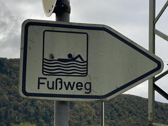 Ein Richtungsschild für einen Fußweg. Darauf ist eine schwimmende Person stilisiert dargestellt.