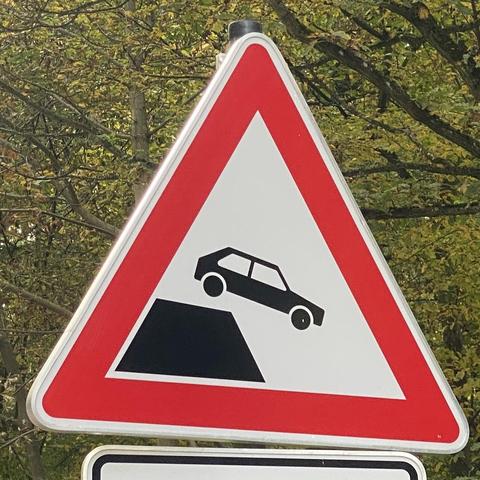 Ein warnendes Verkehrszeichen, das ein Auto darstellt, das von einer Kante oder einem steilen Abhang stürzt, umschlossen von einem roten Dreieck. Im Hintergrund ist unscharfes Blattwerk zu sehen.