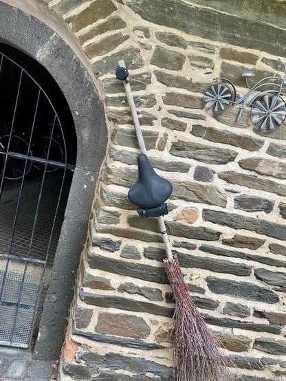 Ein Besen mit einem Fahrradsattel samt Werkzeugtasche und einer Klingel an einer Mauer montiert. Daneben ein kleines metallenes Miniaturfahrrad. 