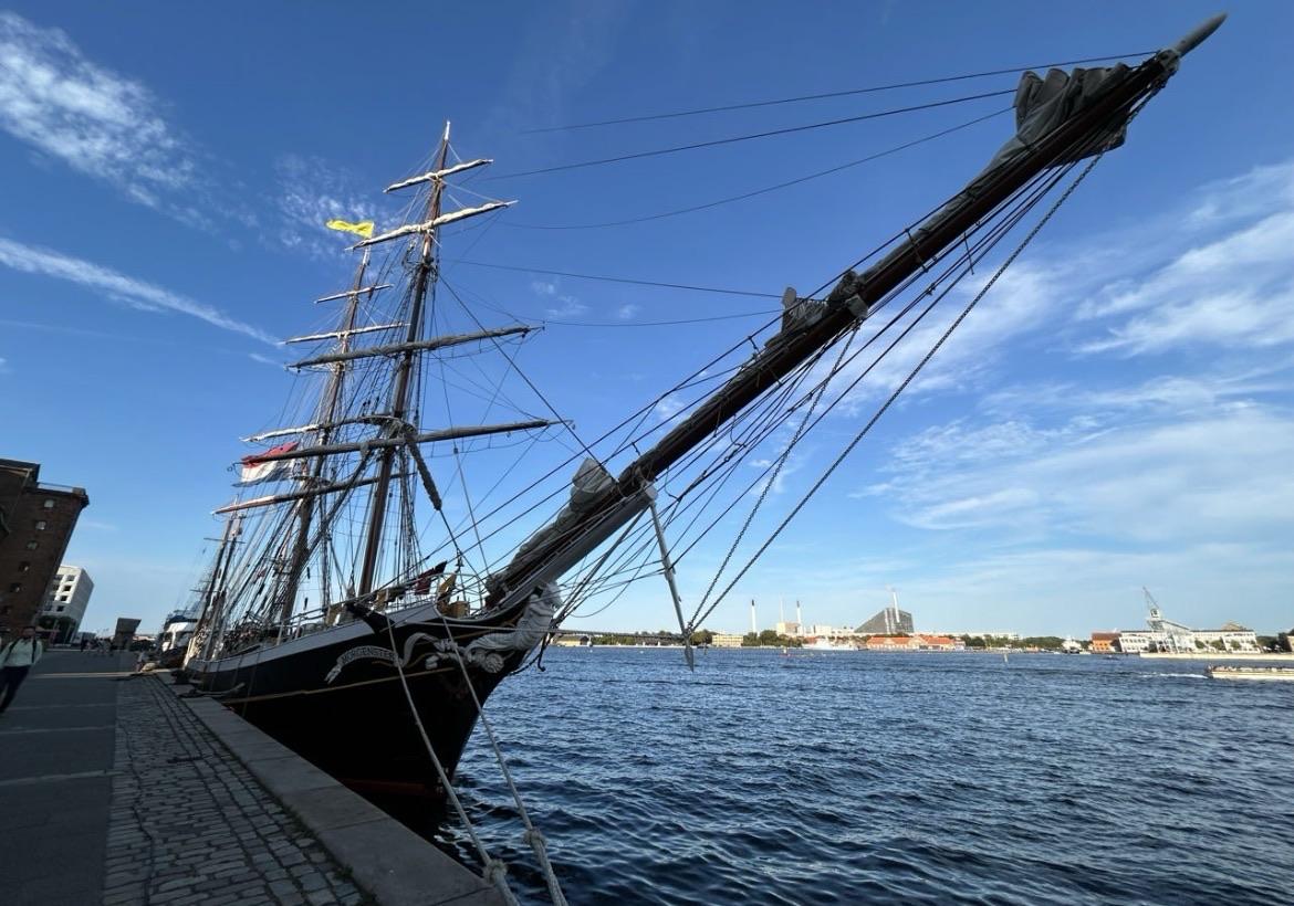 Ein hohes Schiff dockt entlang einer Uferpromenade, wobei sich sein prominenter Mast und seine Takelage in Richtung eines klaren blauen Himmels erstrecken. Im Hintergrund sämen Gebäude das Ufer, und das Wasser spiegelt die Umgebung.


A tall ship is docked along a waterfront, with its prominent mast and rigging extending towards a clear blue sky. In the background, buildings line the shore, and the water reflects the surroundings.
