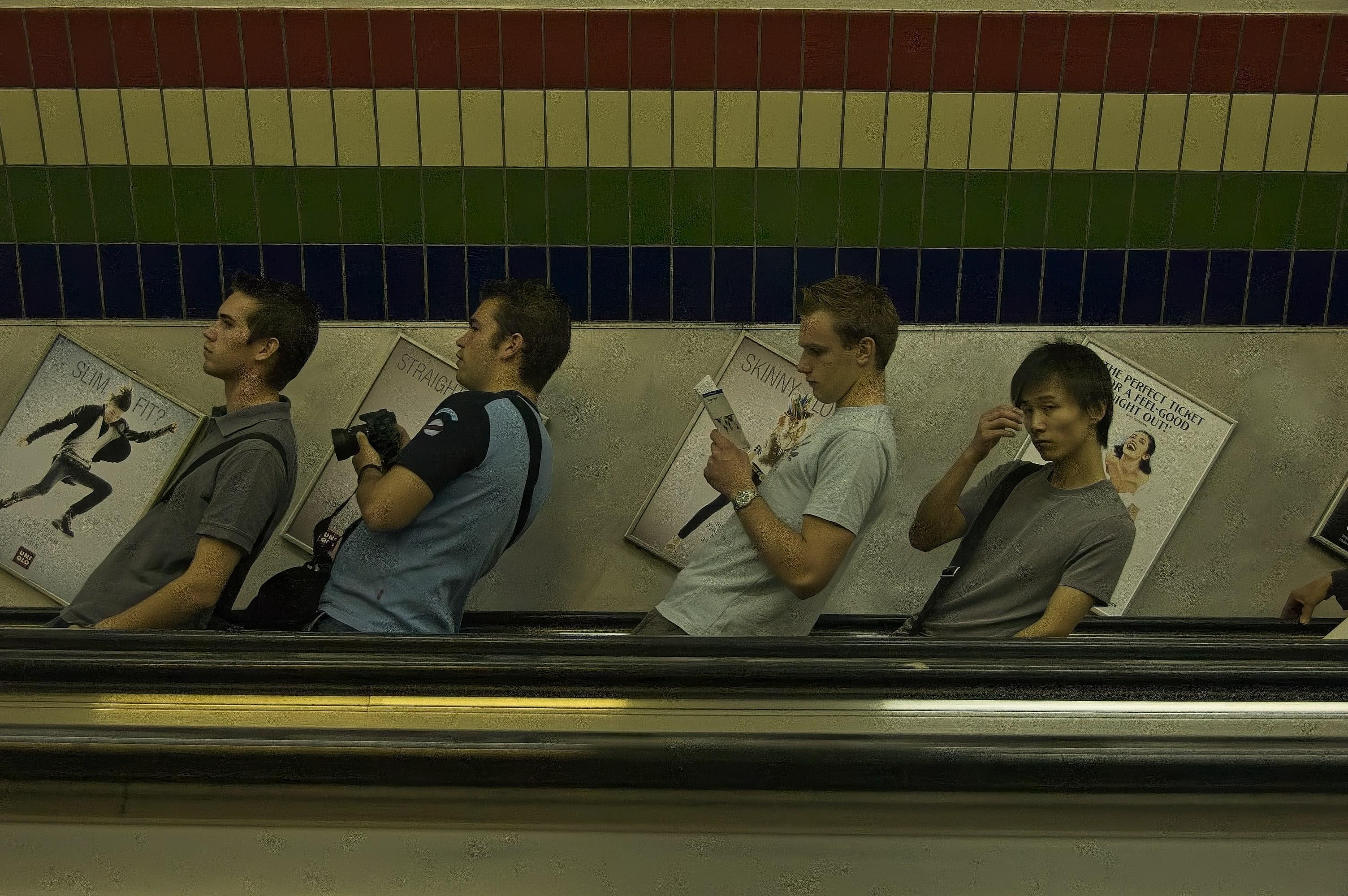 mehrere Menschen auf einer Rolltreppe. Die Personen scheinen schräg zu stehen, da die Kamera auf der schrägen Rolltreppe aufgestellt wurde.