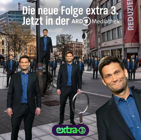Das Bild zeigt einen Mann in einem schwarzen Anzug und einem blauen Hemd, der in einer städtischen Umgebung steht und von mehreren Kopien seiner selbst in einer humorvollen, surrealen Anordnung umgeben ist. Der Text auf dem Bild bewirbt eine neue Episode der Serie "extra 3". 