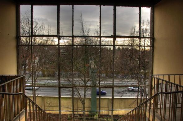 Blick durch ein Fenster ohne Scheiben auf eine Autobahn. Ein Treppengeländer ist zu erfassen. Die Bäume tragen keine Blätter und die Wolken sind grau.