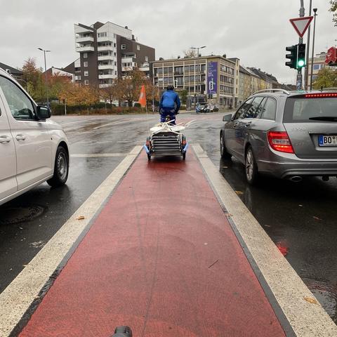 Eine Person in blauer Kleidung fährt mit einem Fahrrad mit Anhänger auf einem roten Radweg. Zwei Autos warten an einer Ampel, und die Umgebung ist regnerisch mit nassem Asphalt. Im Hintergrund sind Gebäude zu sehen.