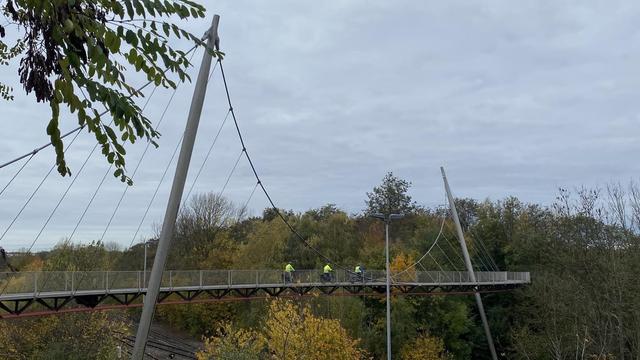 Eine moderne Hängebrücke ist in einer Parklandschaft zu sehen, über die drei Personen in gelben Jacken Rad fahren. Im Hintergrund sind üppige Bäume mit Herbstlaub und ein bewölkter Himmel zu sehen.