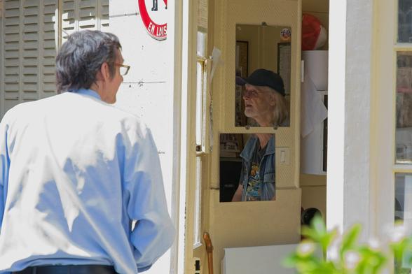Eine Fotoszene mit zwei Personen. Eine Person spricht zu einer Person in einem kleinen Raum. Darin sitzt eine Person, die auf dem Foto auf zwei geteilten Spiegel zu sehen ist.