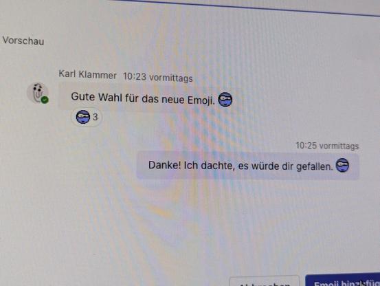 Screenshot von einem Dialog in Microsoft Teams, wo ein Chat simuliert ist. Karl Klammer schreibt "Gute Wahl für das neue Emoji." Daneben ein Mata Smiley, der skeptisch guckt und lila gefärbt ist. Als Antwort steht dort "Danke. Ich dachte, es würde dir gefallen. Daneben steht auch noch Mal der gleiche smiley. Auf die erste Nachricht gibt es drei Reaktionen mit dem Mata Smiley 