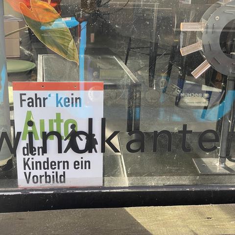 Foto eines Schildes. Auf diesem steht fahr kein Auto den Kindern ein Vorbild. Auf dem Schaufenster steht Windkante