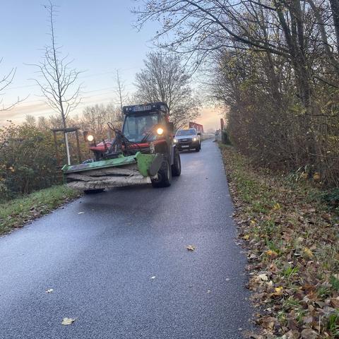 Ein Traktor mit einem großen Bürstenaufsatz fährt auf einem Radweg, die von Bäumen und Laub gesäumt ist. 