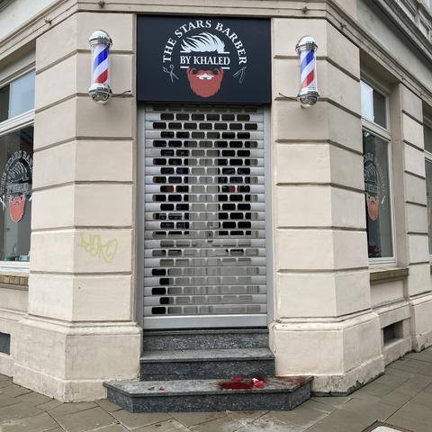 Bild der Außenansicht eines Friseursalons mit einem Schild mit der Aufschrift „THE STARS BARBER BY KHALED“. Der Eingang ist mit einem Metallgitter gesichert, und auf beiden Seiten befinden sich Friseurstangen. Auf dem Boden sind rote Blutlachen zu sehen, die möglicherweise auf einen Unfall hinweisen. 