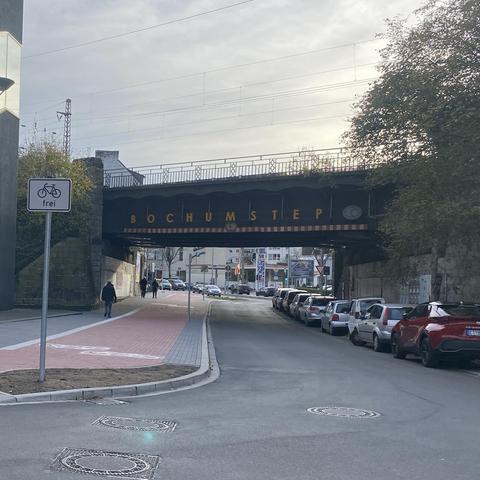 Eine Straßenansicht mit einer Eisenbahnbrücke mit der Aufschrift „BOCHUMSTEPS“ darüber. Darunter befindet sich ein Fahrradwegschild, das darauf hinweist, dass dieser Gehweg mit dem Rad benutzt werden kann, sowie Fußgänger, die auf dem Gehweg gehen. Mehrere geparkte Autos säumen die Straße, mit einer städtischen Kulisse aus Gebäuden im Hintergrund.