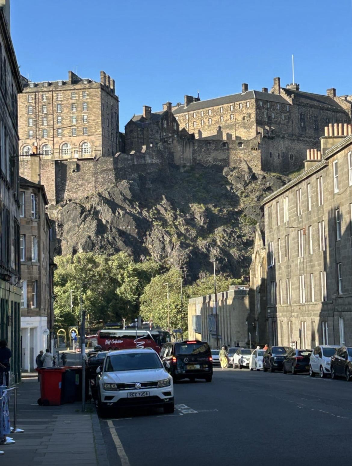 Eine Straßenansicht mit historischen Steingebäuden, einer Reihe von geparkten Autos und einem klaren blauen Himmel. Im Hintergrund thront ein Schloss auf einem felsigen Hügel, was darauf hindeutet, dass sich Edinburgh befindet.


A street view featuring historic stone buildings, a line of parked cars, and a clear blue sky. In the background, a castle is perched on a rocky hilltop, suggesting the location is Edinburgh.
