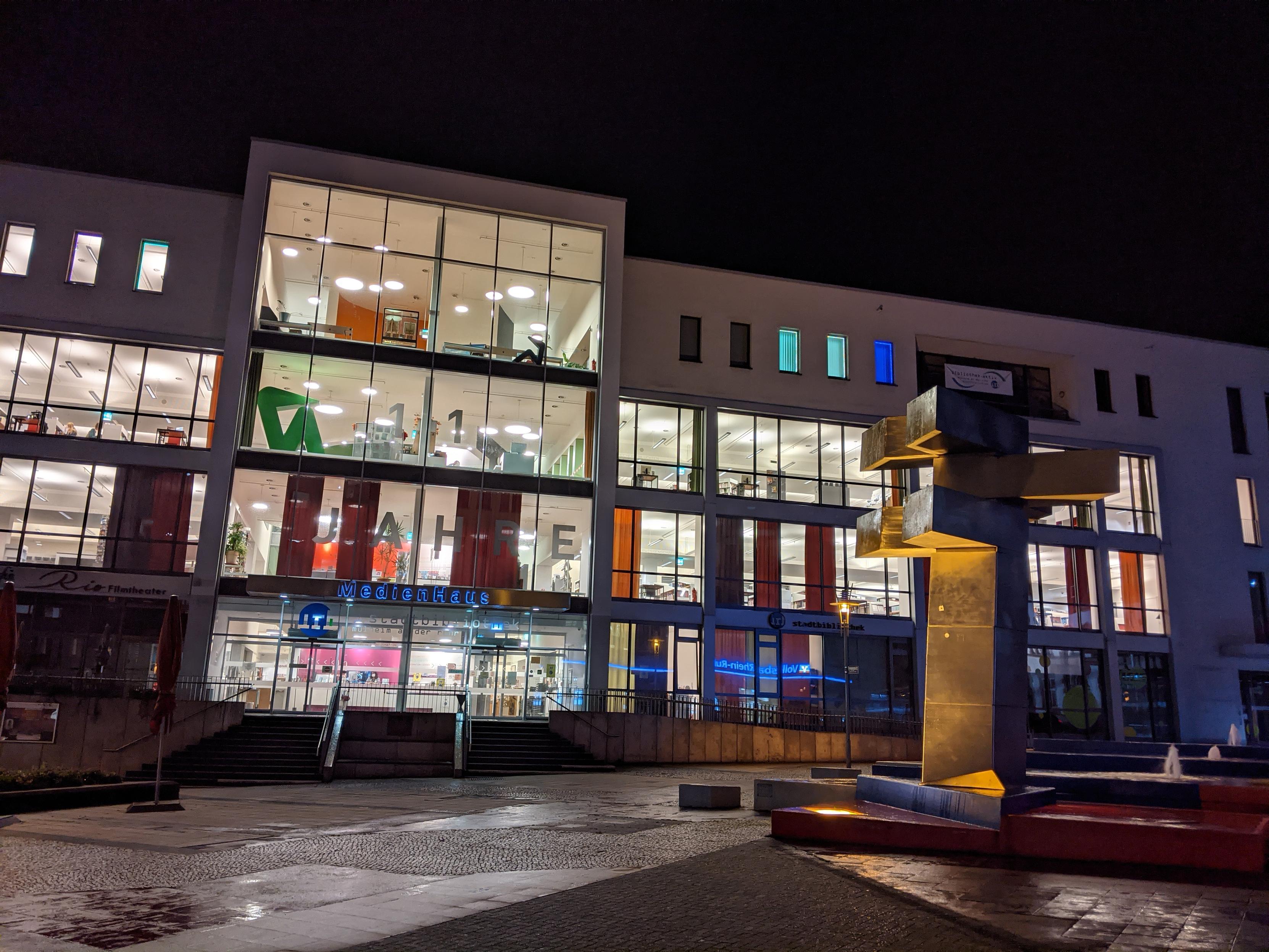Eine nächtliche Außenansicht des Medienhauses in Mülheim an der Ruhr. Das moderne Gebäude hat große Glasfenster und ist hell erleuchtet. Davor steht die silberne, abstrakte Skulptur des Hajek-Brunnens. Die Skulptur wird von unten beleuchtet, was Teile davon in Gelb/Gold erscheinen lässt. Der Brunnen und der Platz davor sind nass und spiegeln die Lichter wider. Im Zentrum des Medienhauses ist in der 2. Etage die Zahl 11 und in der ersten Etage das Wort Jahre in den beleuchteten Fenstern erkennbar.