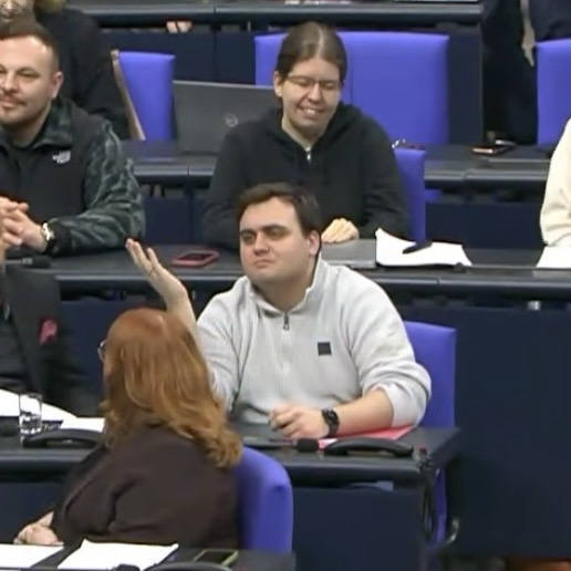 Ein Typ (ich) sitzt im Parlamentssaal des Bundestags, hebt ratlos die Hand und fragt sich offenbar, wie er hier eigentlich gelandet ist – während andere Personen hinter ihm amüsiert lächeln.