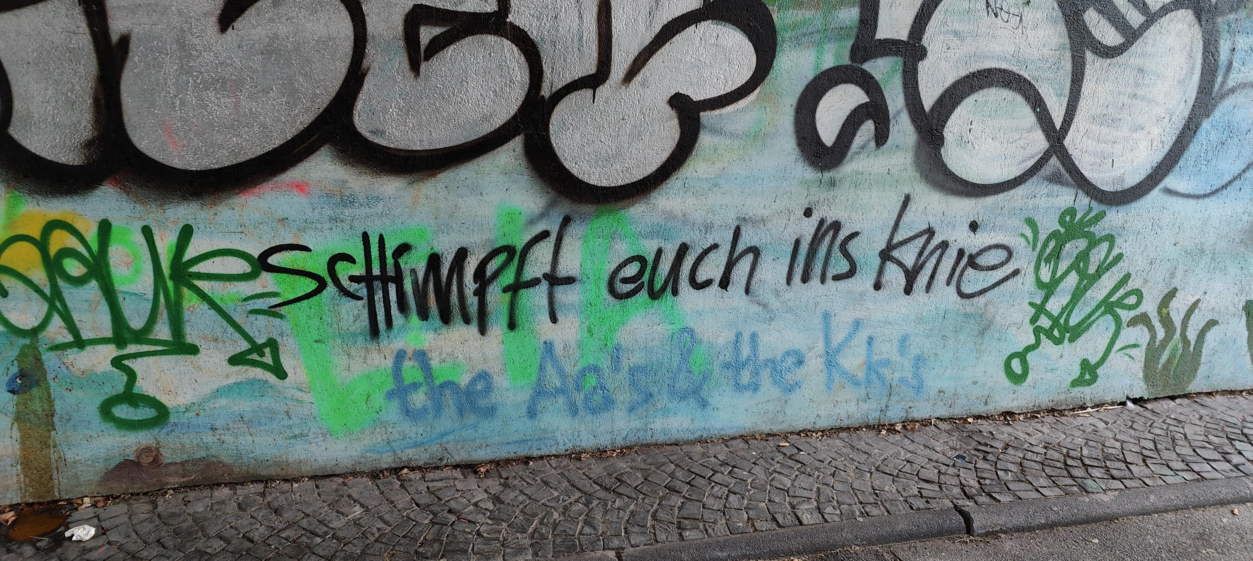 Bunt besprühte Wand auf der mit schwarzer Sprayfarbe der Text schimpft euch ins Knie steht.