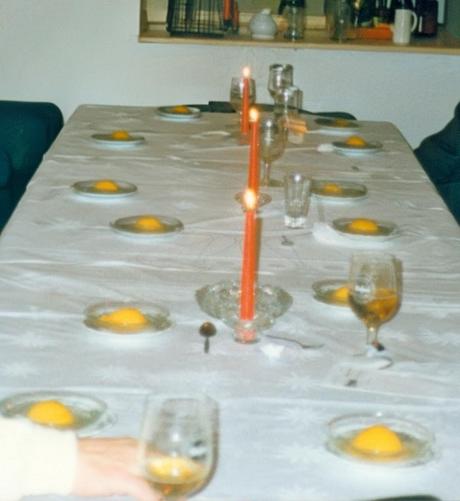 Foto eines Tisches im Restaurant in 1989.
Neben drei roten Kerzen und einigen Biergläsern sind 11 flache, sehr schlichte Glasteller zu sehen, auf denen jeweils nur ein einziger, halber Dosenpfirsich liegt, ohne jegliche weitere Beilage oder Dekoration. 