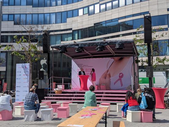 Foto einer Bühne vor dem Kö-Bogen. Zwei Frauen stehen auf der Bühne, Zuschauende sitzen davor. Das Wetter ist sonnig. I'm Hintergrund ist Werbung für den Tag zu sehen, mit einer pinken Schleife