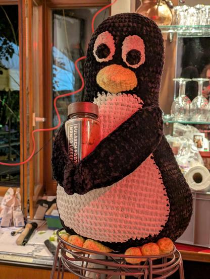 Ein gestrickter Pinguin (Tux) hält eine Gewürzdose mit der Aufschrift "Gulasch fad? MRMCD liefert!". Hintergrund besteht aus Gläsern einer Bar und einem offenen Fenster