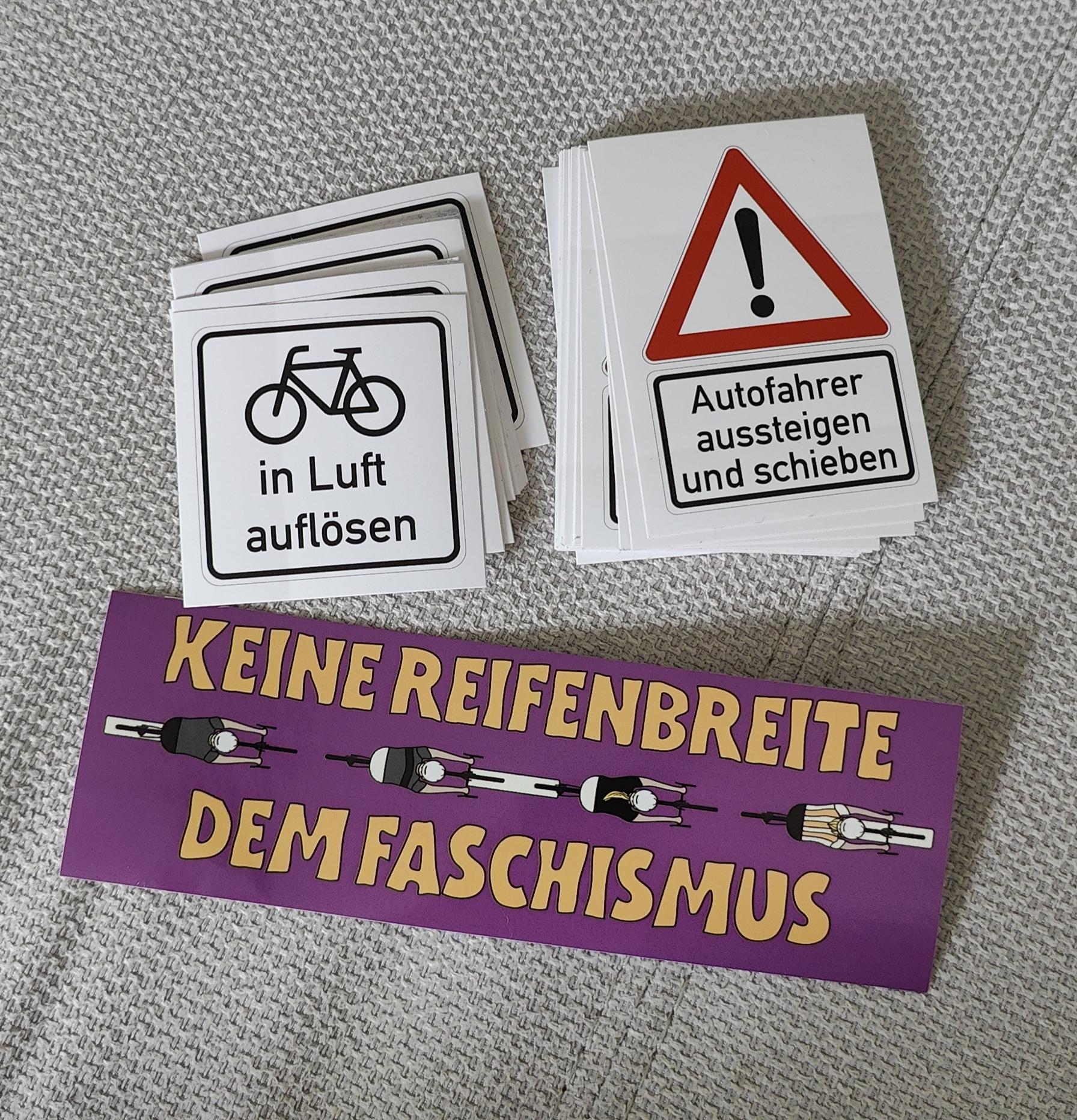 Drei verschiedene Sticker. Zwei sind Verkehrsschildern nachempfunden, auf einem steht ein Fahrradsymbol und darunter der Text in Luft auflösen. Auf dem anderen ist ein Gefahrenschild und darunter der Zusatz Autofahrer aussteigen und schieben.
Der dritte Sticker ist lila und zeigt vier Radfahrende von oben dazu der Text keine Reifenbreite dem Faschismus